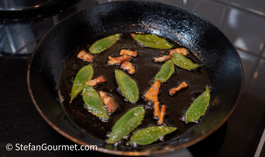 A skillet with sizzling bacon pieces and bay leaves in hot oil.