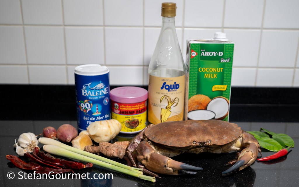 An assortment of ingredients for cooking, including a crab, coconut milk, fish sauce, shrimp paste, fresh herbs, garlic, shallots, chilies, lemongrass, and turmeric laid out on a black countertop.