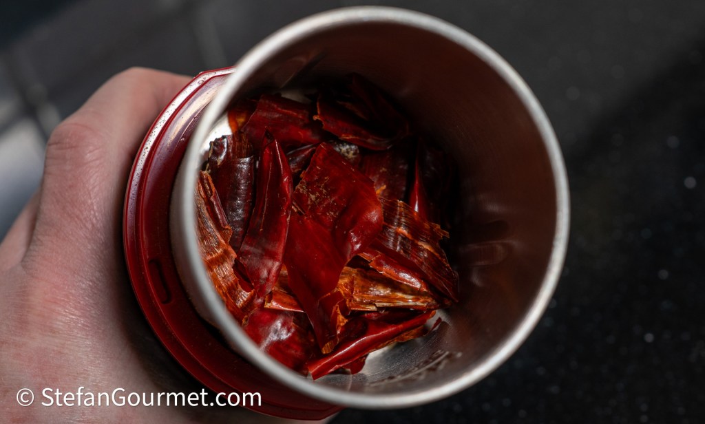 A close-up view of dried red chili peppers inside a metal container held in a hand.