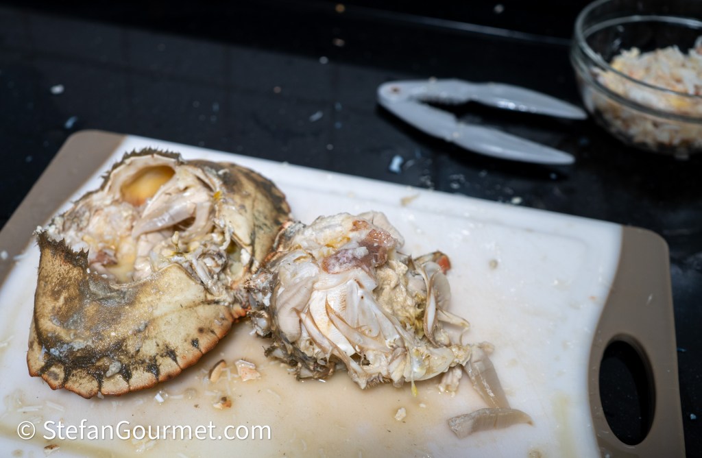 A freshly cracked crab on a cutting board, displaying its shell and crab meat, with a bowl of shredded crab meat and a crab cracker in the background.