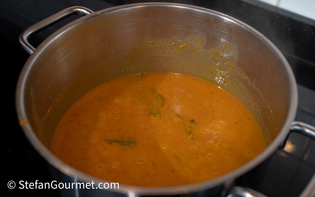A stainless steel pot filled with orange curry simmering on the stove, with visible herbs and spices.