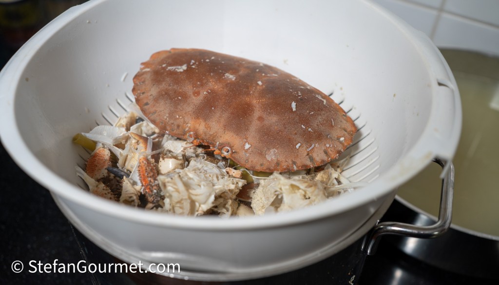 A close-up view of a colander containing cooked crab remains, including a large crab shell and pieces of crab meat.