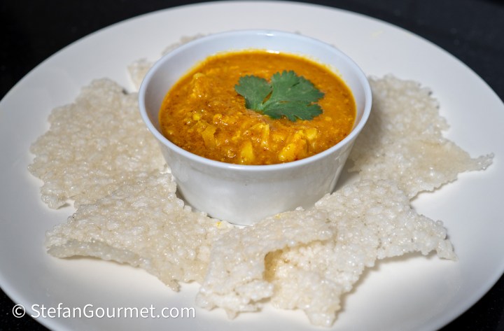 A bowl of curry topped with a cilantro leaf, served with crispy rice crackers arranged around it on a white plate.
