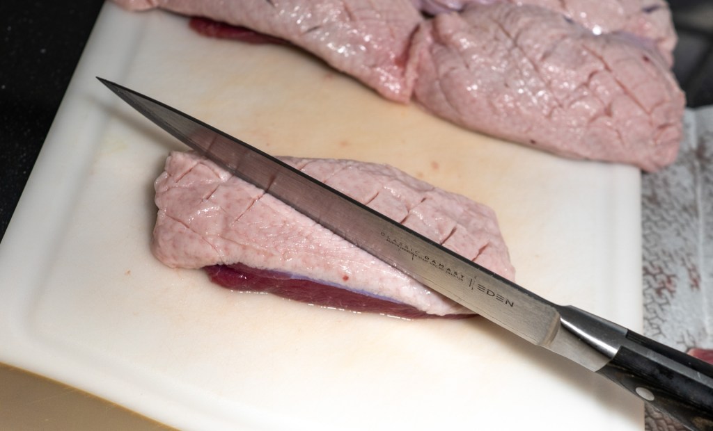 A sharp knife cutting through a piece of raw meat on a white cutting board, with additional meat pieces in the background.
