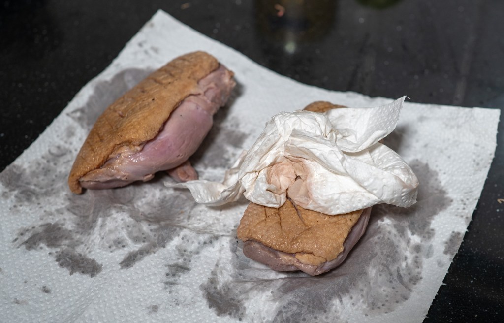 A close-up image of two pieces of raw meat, partially covered with a paper towel, resting on a paper towel on a dark surface.
