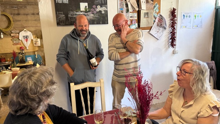 Four people engaged in conversation around a table with glasses of wine, one person holding a bottle of wine, in a cozy indoor setting.