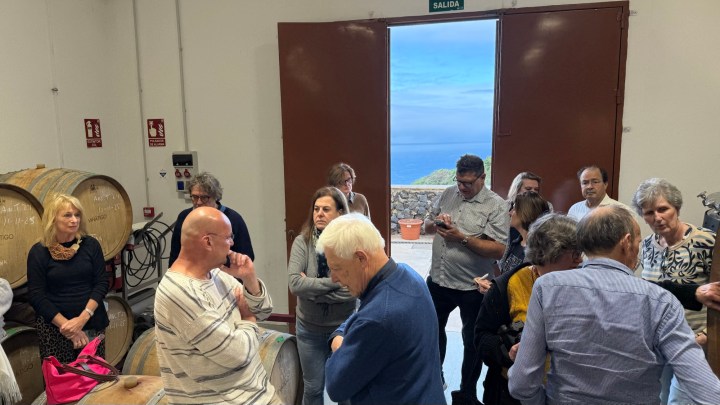 A group of people gathered inside a winery, some sitting on barrels and others standing, with a view of the ocean visible through an open door.