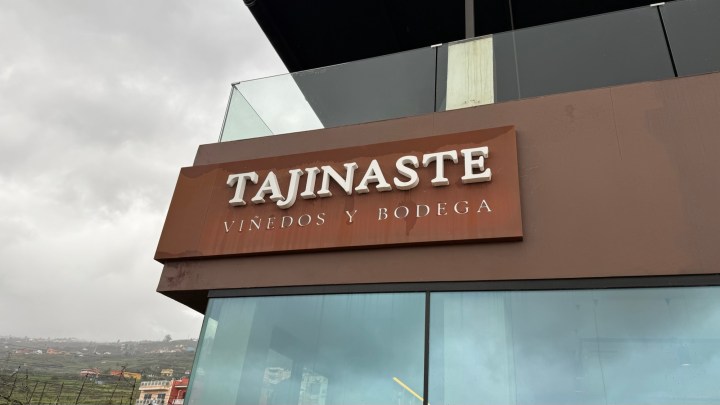 Sign displaying the name 'Tajinate' and the text 'Viñedos y Bodega' on a modern building with a cloudy sky in the background.