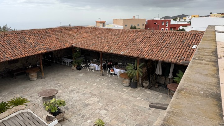 A view of a rustic courtyard featuring a terracotta roof, stone flooring, and tables with chairs set up for dining. Potted plants and palm trees are visible around the area. In the background, colorful buildings are present under a cloudy sky.