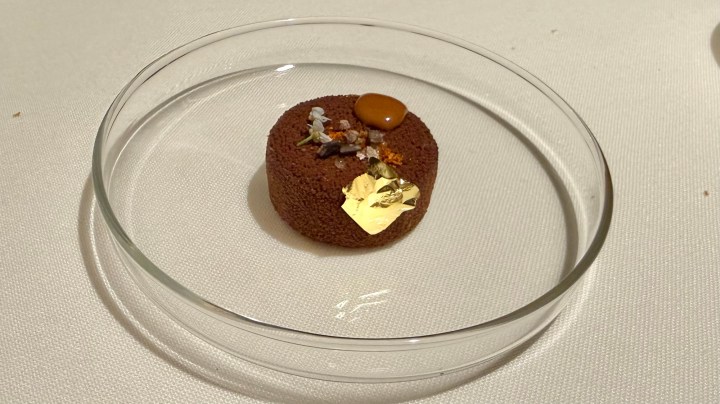 A beautifully plated chocolate dessert featuring a round, textured chocolate cake topped with a golden leaf, a drizzle of caramel, and garnished with small flowers and crystals, served in a clear glass dish.