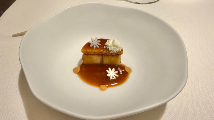 A gourmet dessert presented on a white plate, featuring layers of cake or pastry drizzled with a glossy caramel sauce, garnished with small edible flowers and a dusting of powdered sugar.
