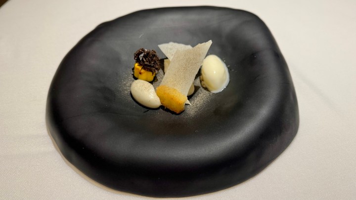 A sleek black plate featuring an artistic dessert arrangement, including various textures and colors like a crumbled topping, cream dollops, and thin crispy sheets.