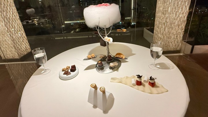 A beautifully presented dessert table featuring various intricate sweets, a decorative cotton candy display on a stand, and two glasses of water, all set on a white tablecloth with a city view in the background.