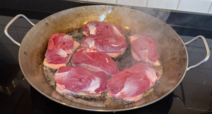 Raw meat sizzling in a large frying pan on a stovetop, surrounded by steam.