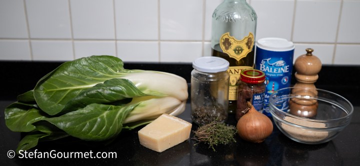 A collection of cooking ingredients including bok choy, cheese, spices, a bottle of alcohol, a jar of herbs, sugar, salt, and an onion on a black countertop.