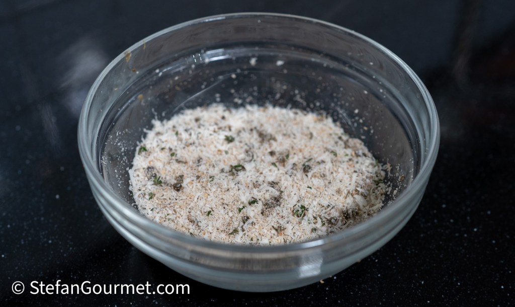 A glass bowl filled with a mixture of breadcrumbs and herbs resting on a black surface.