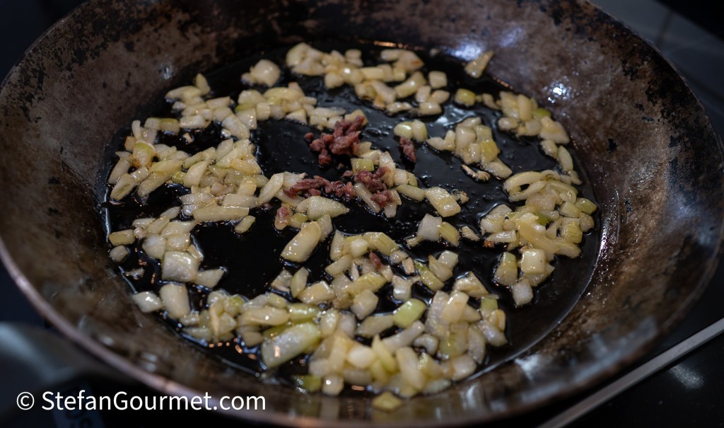 Close-up of a frying pan with diced onions and small pieces of bacon cooking in dark oil.