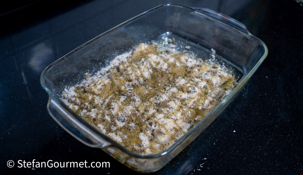 A glass baking dish filled with layered pasta topped with breadcrumbs and herbs, placed on a dark countertop.