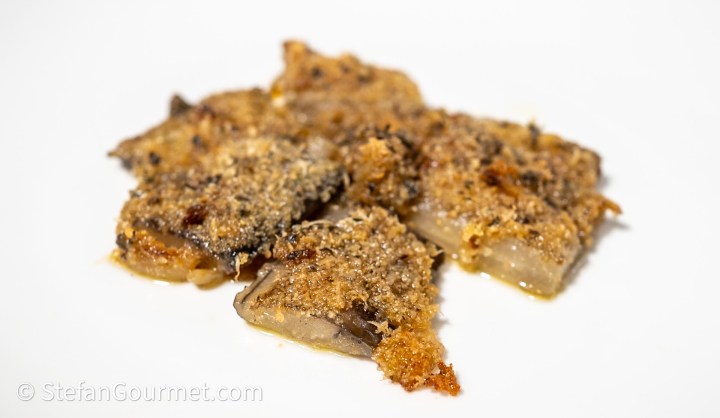 A close-up of several pieces of baked or fried fish, coated in a crispy, golden-brown breadcrumb topping, arranged on a white plate.