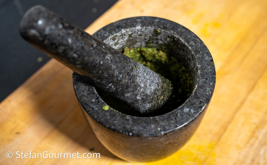 A close-up of a dark stone mortar and pestle with green mixture inside, positioned on a wooden surface.