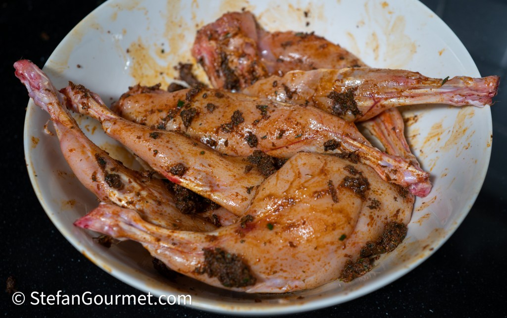 A bowl containing marinated rabbit legs coated in spices and herbs.