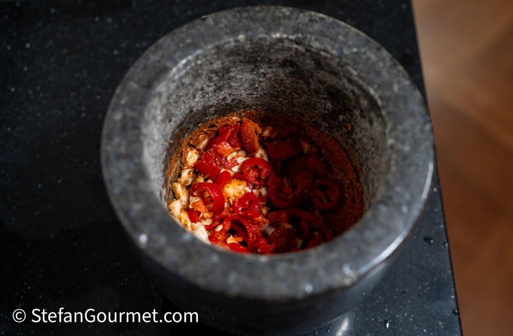 A granite mortar filled with chopped red peppers and spices, placed on a dark countertop.