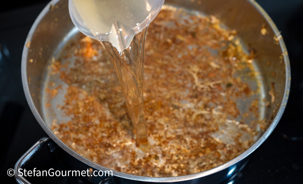 Pouring liquid into a pan with browned bits on the bottom, indicating preparation for making a sauce.