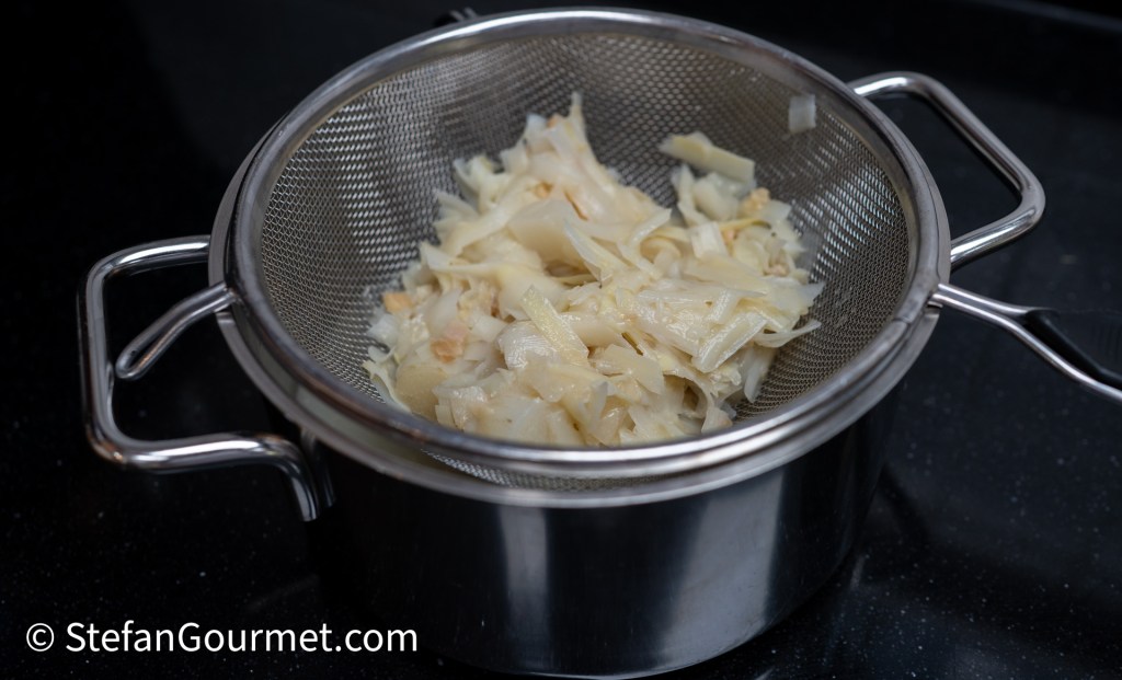A stainless steel colander filled with drained cooked pasta or noodles, sitting on a dark countertop.