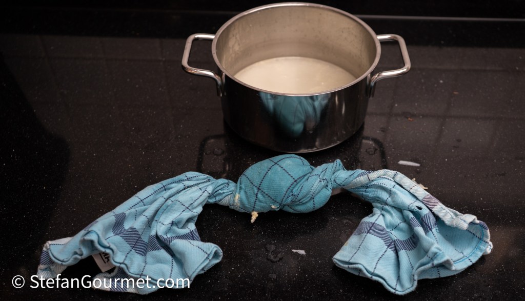 A stainless steel pot filled with a white liquid sits on a black countertop, accompanied by two blue checkered kitchen towels, one twisted.