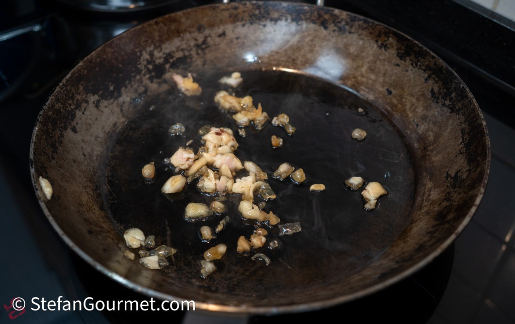 Sizzling garlic and small pieces of meat frying in a dark, slightly worn frying pan on a stovetop.