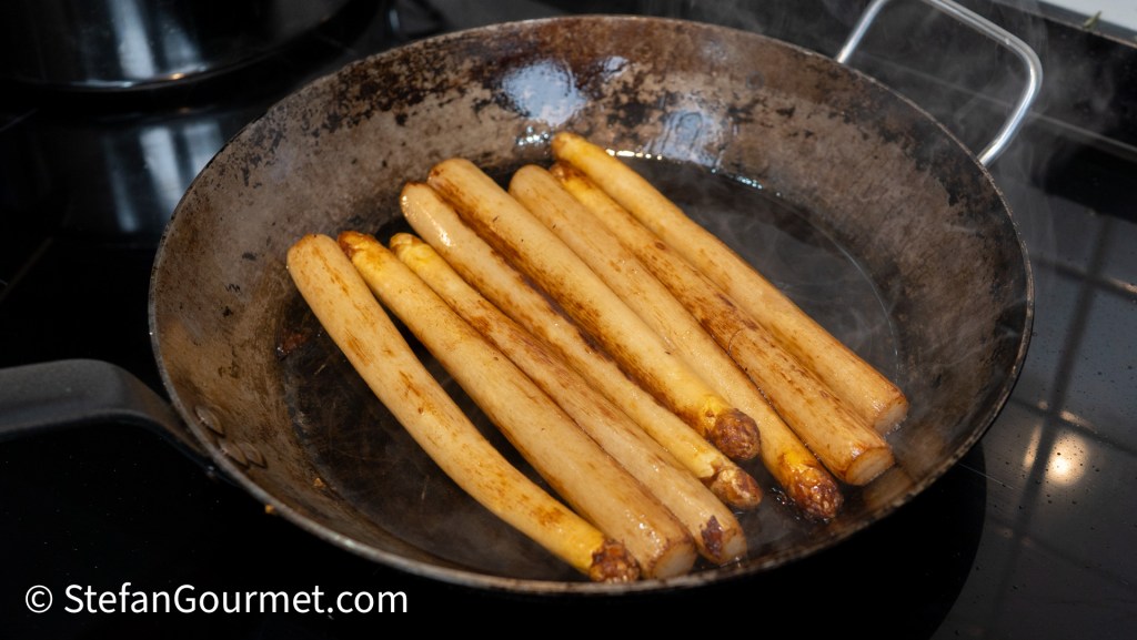 A cast iron skillet with several long, golden-brown cooked vegetables sizzling in a light steam, placed on a stovetop.