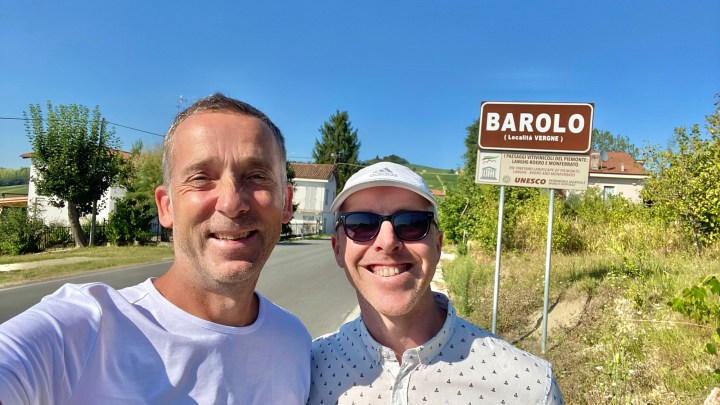 Two smiling men posing for a selfie in front of a sign that reads 'Barolo,' indicating the location. The background features greenery and a clear blue sky.