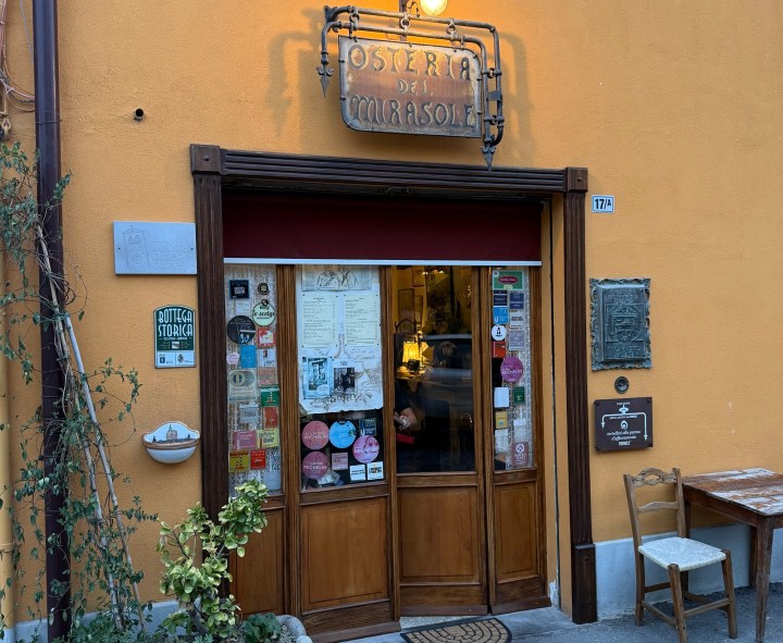 Front view of 'Osteria dei Mirasole' with wooden door, colorful stickers, and signage on a yellow wall.