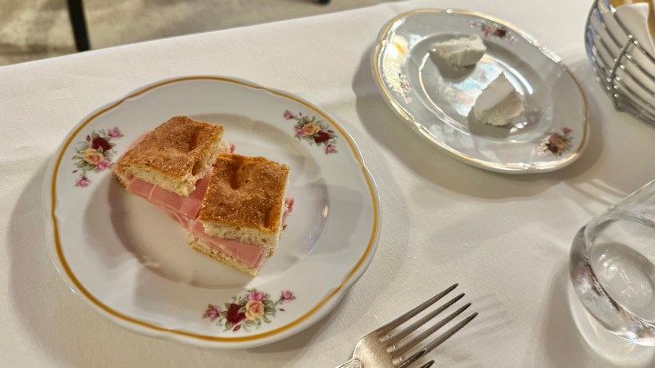 A slice of ham sandwich on a decorative plate, with a plate of two pieces of cheese in the background.