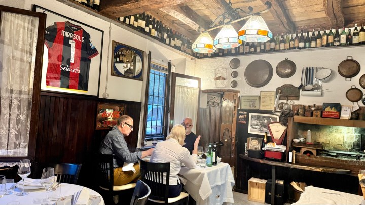 Interior of a cozy restaurant featuring wooden decor, a vintage jersey on the wall, and a display of wine bottles overhead. Two patrons are seated at a table, engaging in conversation.
