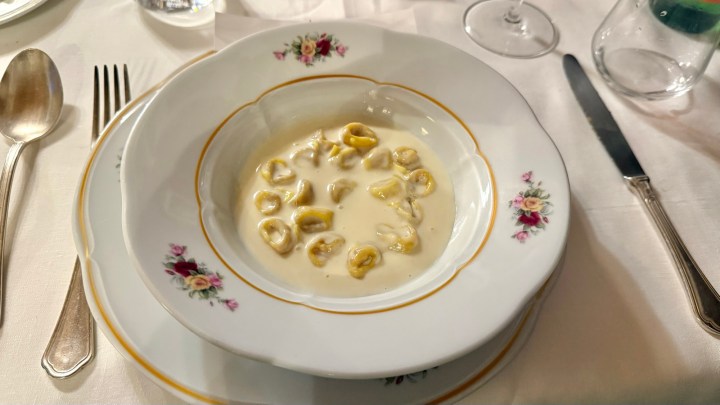 A decorative white bowl filled with yellow tortellini in a creamy sauce, placed on a floral-patterned plate, with silverware and a glass in the background.