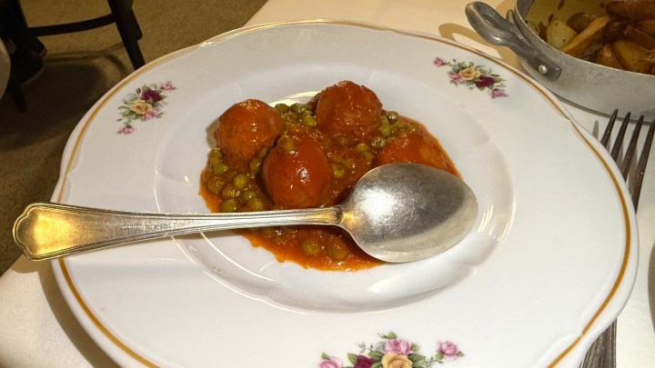 A plate of meatballs in a tomato sauce with green peas, served with a large silver spoon on a decorative white plate.