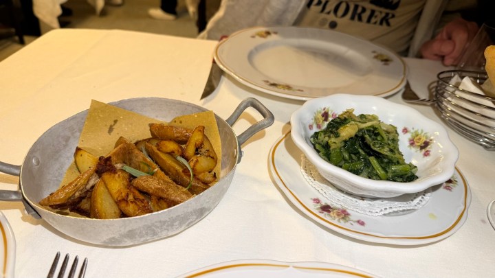 A dish of golden, crispy roasted potatoes served in a metal pan alongside a bowl of sautéed greens.