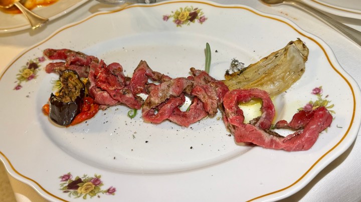 A plate featuring thinly sliced cooked beef arranged in a wavy pattern, accompanied by a piece of grilled onion and roasted vegetables, garnished with black pepper.
