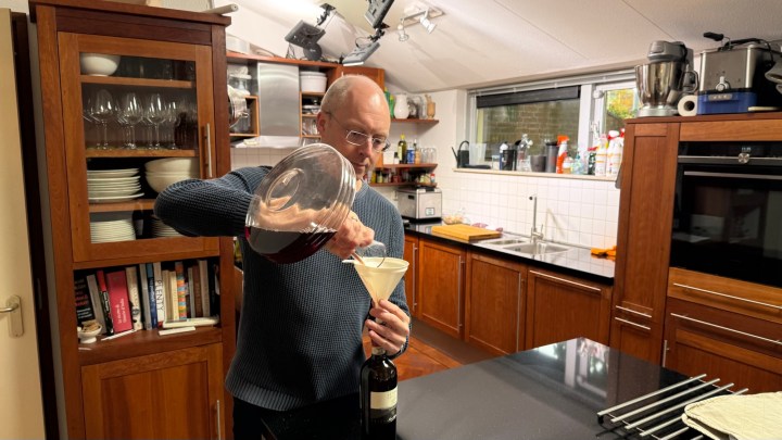 A person pouring liquid from a large container into a funnel, positioned above a wine bottle, in a modern kitchen setting.