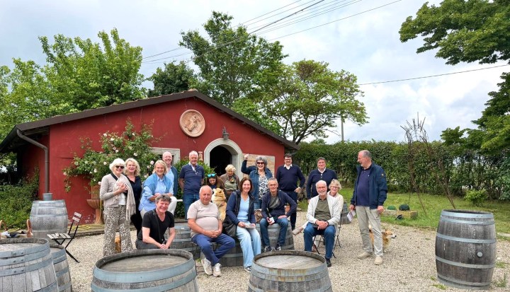A group of people gathered outdoors in front of a red building, surrounded by greenery and wooden barrels.