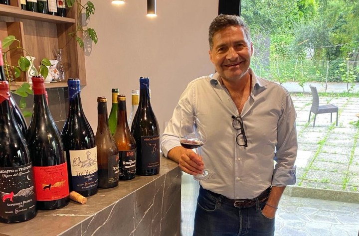 A man holding a glass of wine stands next to a variety of wine bottles on a counter, with a natural setting visible through a window in the background.