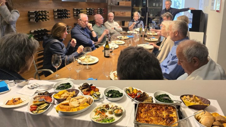 A group of people sitting at a long table enjoying a meal with wine in a cozy dining setting, featuring a variety of dishes including vegetables, bread, and a baked casserole.