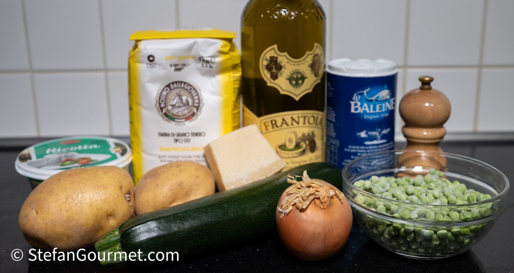 A collection of fresh ingredients including potatoes, zucchini, an onion, green peas, a block of cheese, ricotta, flour, olive oil, and salt in a wooden grinder, arranged on a black countertop.