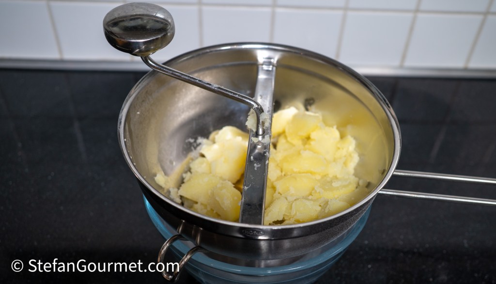A stainless steel food mill containing mashed potatoes, with a hand crank and a blue bowl beneath it, sitting on a black countertop.