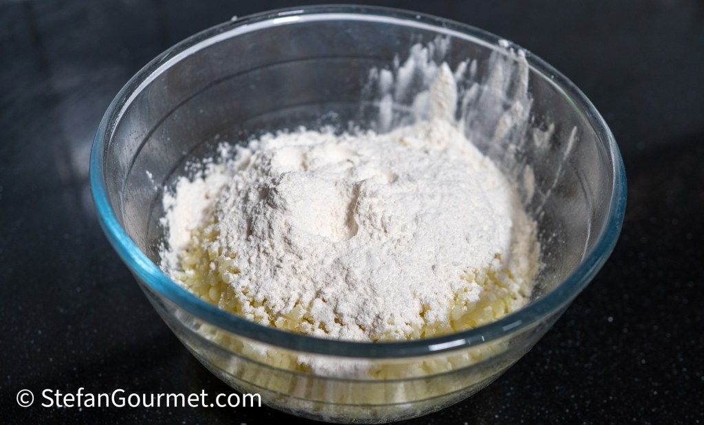 A clear glass bowl containing a mix of butter and flour on a dark countertop.