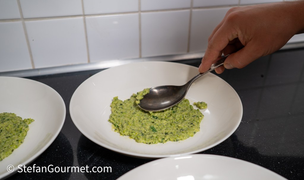 A hand using a spoon to serve a green puree onto a white plate, with additional plates of similar puree in the background.