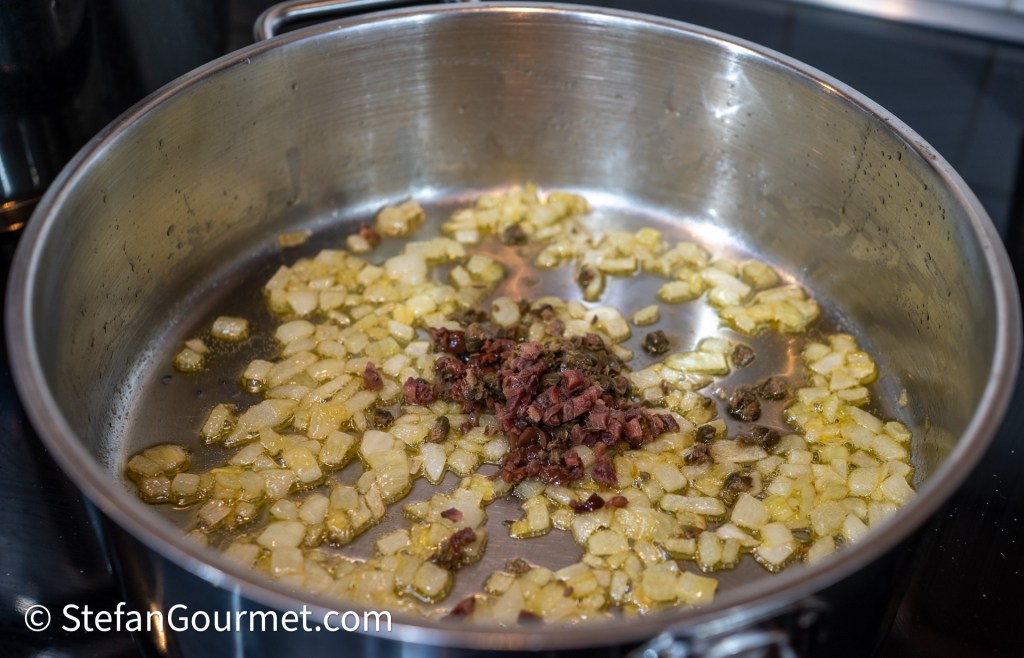 Sautéed onions and chopped ingredients in a stainless steel pan with olive oil on a stovetop.
