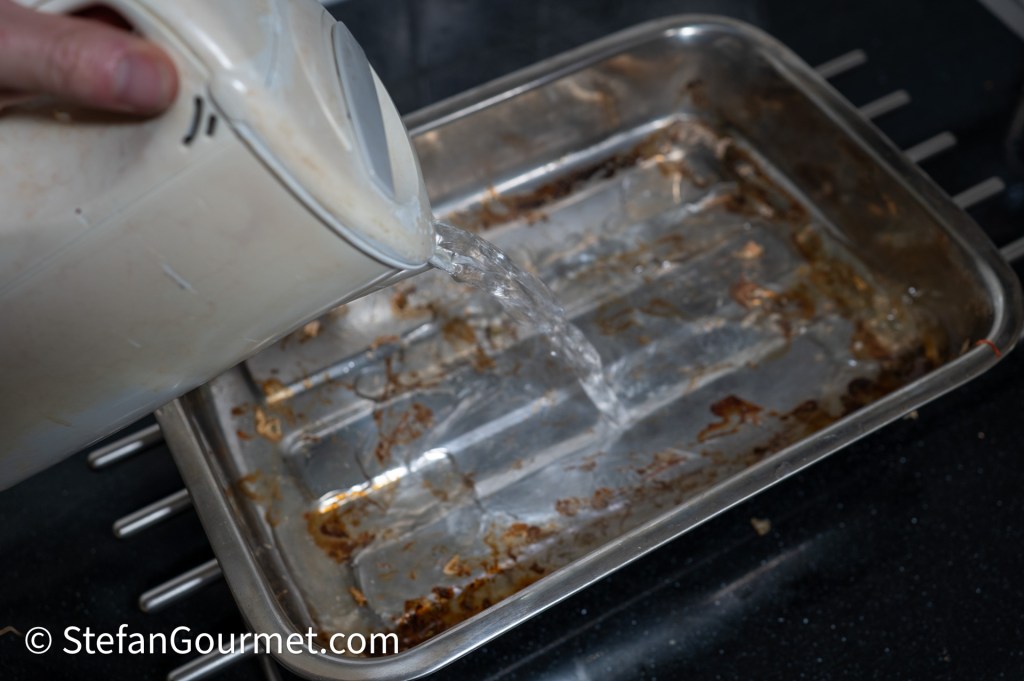 A person pouring water from a container into a dirty metal baking pan on a black countertop.