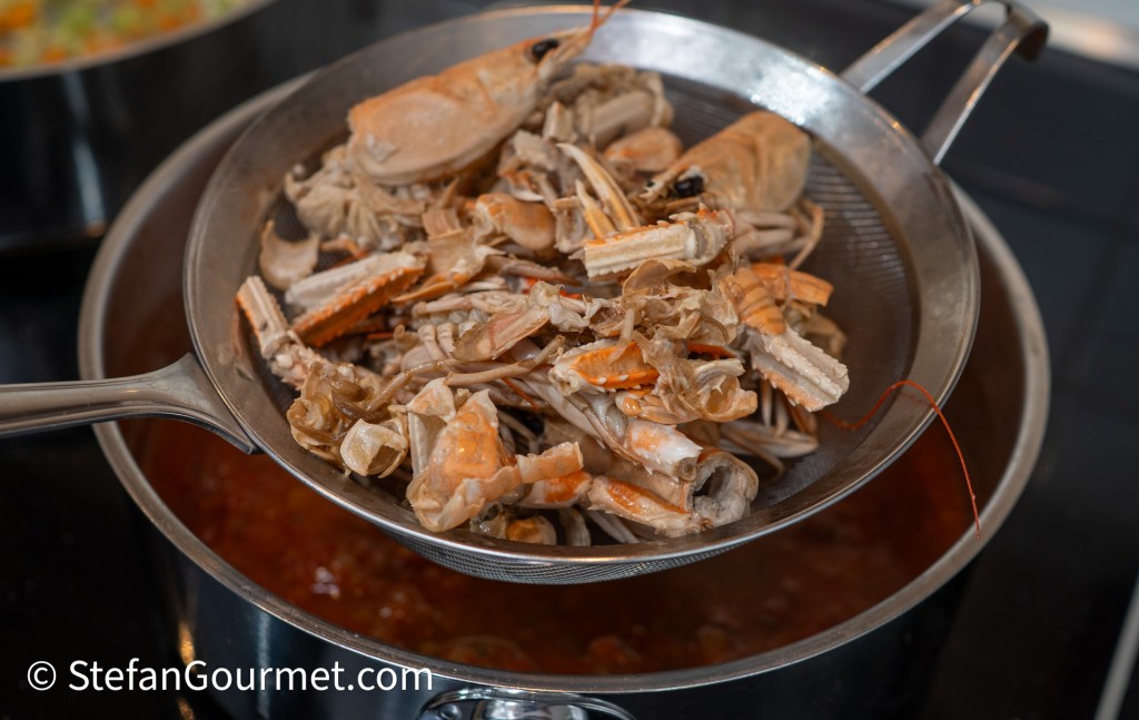A metal strainer holding cooked seafood shells above a pot of simmering sauce.